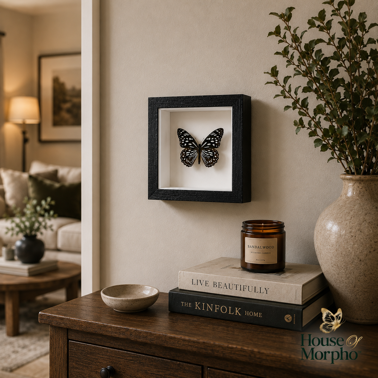 A black square shadow box frame mounted on a beige wall, containing a black and white butterfly specimen. Below the frame, a wooden console table holds a candle, books, and a ceramic vase with greenery in a styled living room setting.