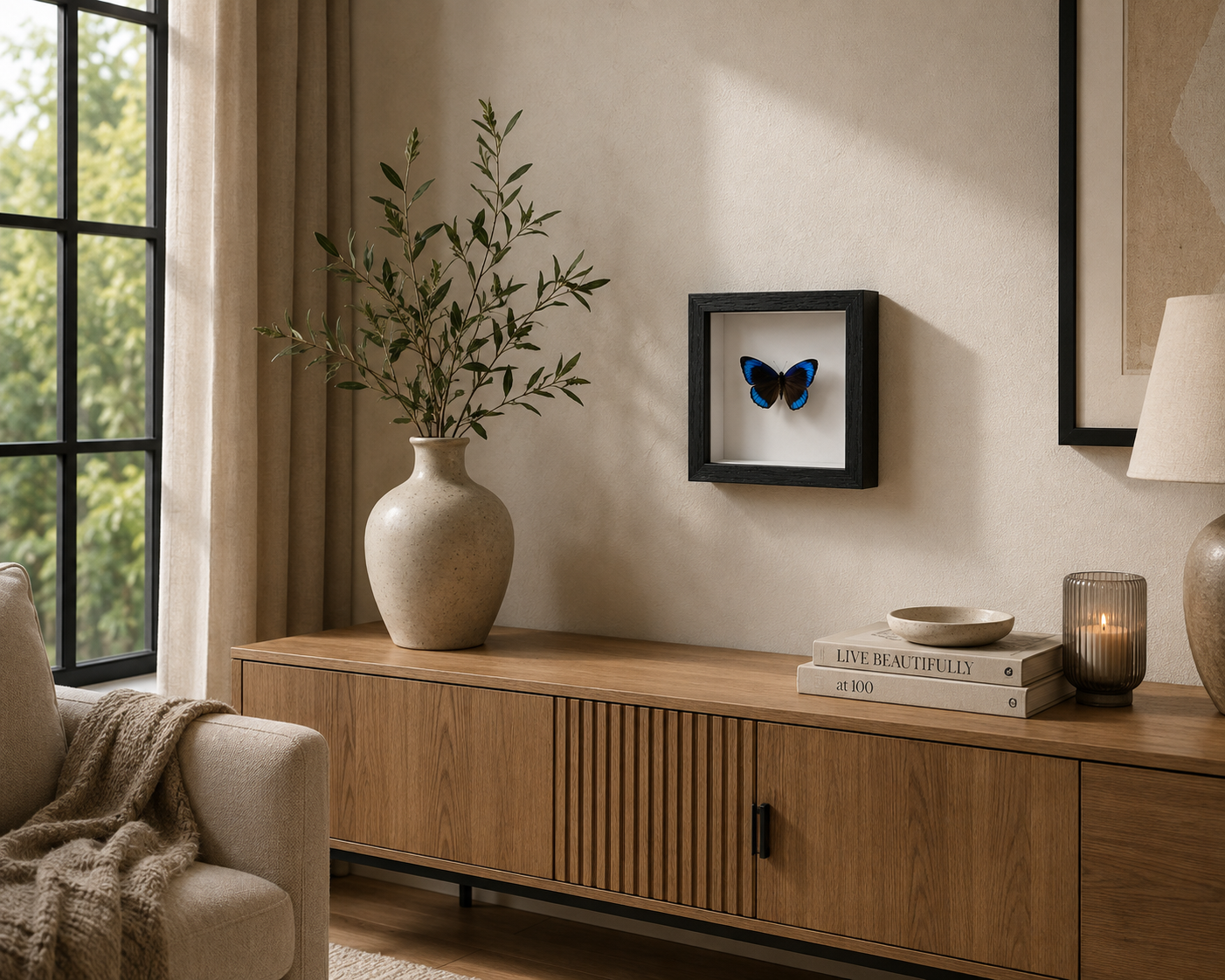 A framed butterfly wall art piece hangs on a beige wall above a wooden console table in a styled living room. The black-framed shadow box contains a blue and black butterfly, with a beige vase, books, and a candle nearby.