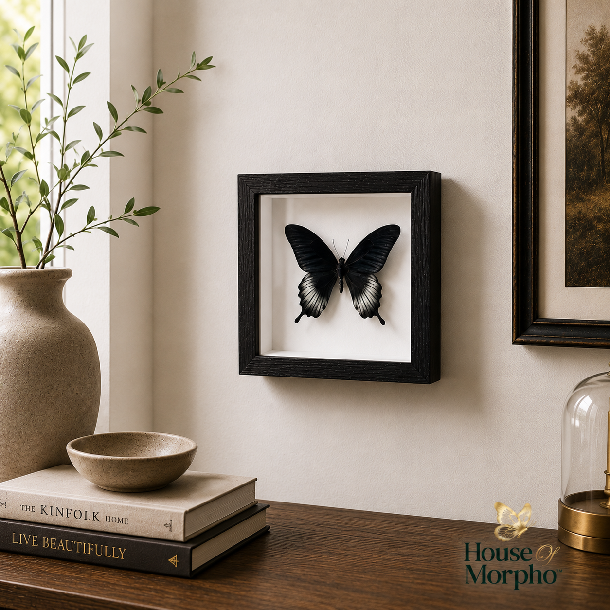A black square frame displaying a black and white butterfly mounted on a white backing, hung on a beige wall above a wooden table with books, a vase, and a bowl.