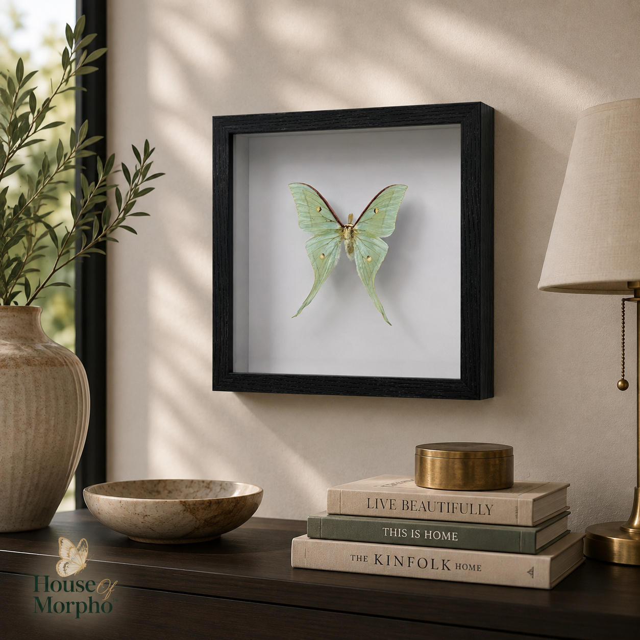 A framed butterfly wall art piece hangs on a beige wall above a dark wooden console table. The frame contains a light green butterfly mounted on a white background, surrounded by decorative props including a vase with greenery, a bowl, stacked books, and a table lamp.