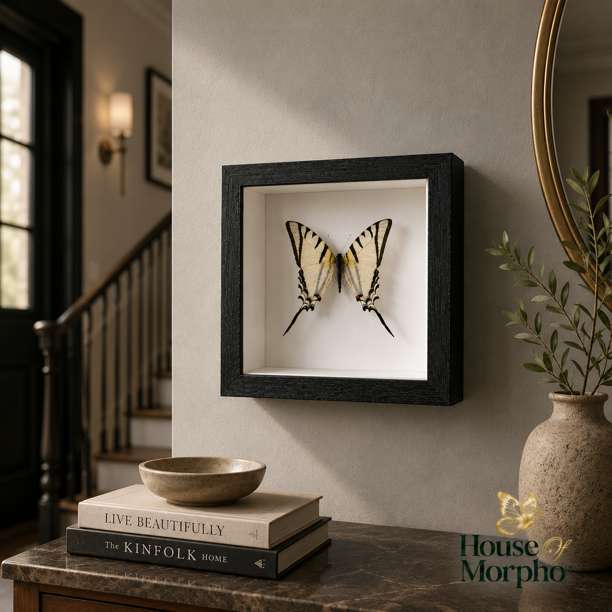 A black shadow box frame displaying a pair of yellow and black tiger swallowtail butterflies mounted on a white backing, hung on a beige wall in a styled home interior setting.