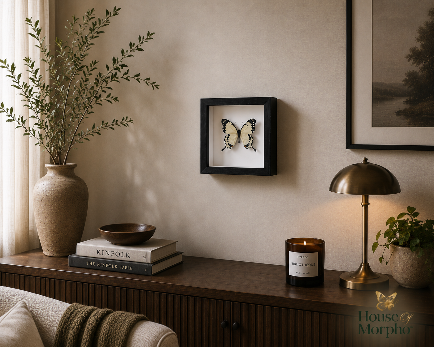 A framed butterfly taxidermy display hangs on a beige wall above a wooden console table styled with a vase, books, a lamp, and a candle in a warm, elegant room setting.