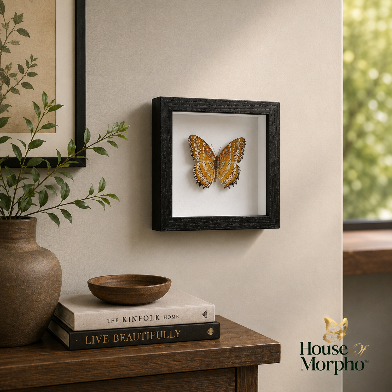 A framed butterfly taxidermy wall art piece displayed on a beige wall above a wooden console table with books and a plant. The black frame contains a yellow and brown butterfly mounted on a white background.