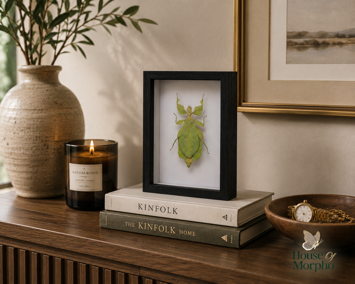 A framed green leaf-shaped insect taxidermy specimen displayed on a stack of books on a wooden console table, styled with a lit candle, a ceramic vase with greenery, and a gold-framed picture on a beige wall.