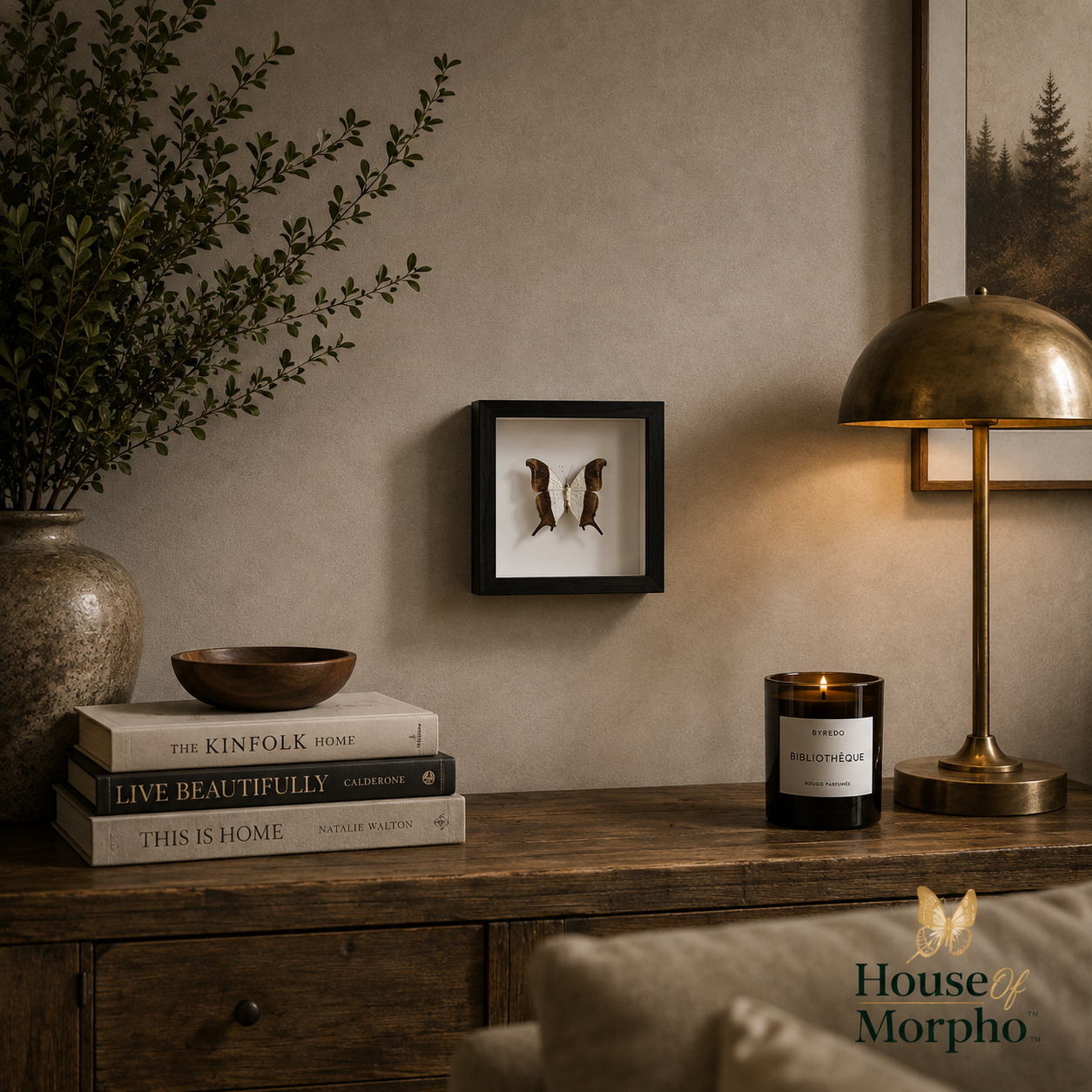 A framed wall art piece featuring a pair of brown boots mounted on a beige wall above a wooden console table. The scene is styled with a brass table lamp, a black candle jar, a stack of books, and a large vase with greenery.