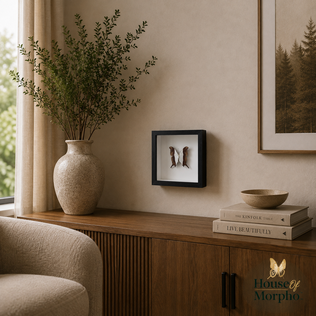 A framed butterfly wall art piece in a black frame hangs on a beige wall above a wooden console table. The scene includes a large beige ceramic vase with greenery, a small bowl, stacked books, and part of a chair, in a styled interior setting.