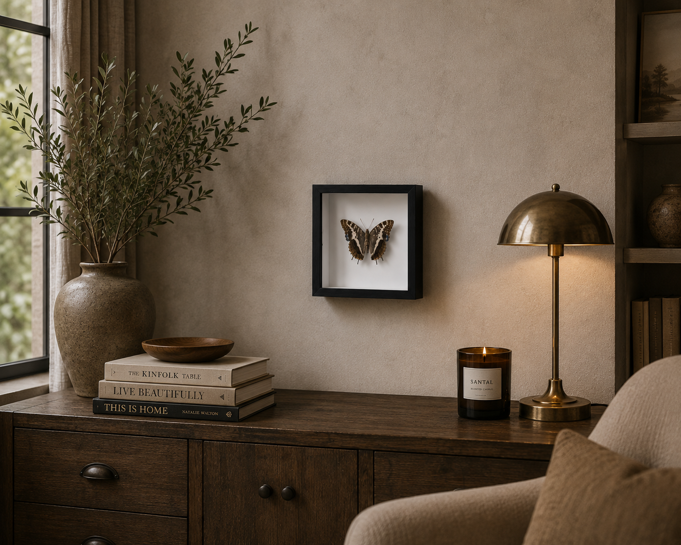 A framed butterfly specimen display hangs on a textured beige wall above a dark wood console table in a styled room setting with a brass table lamp, books, and a vase.